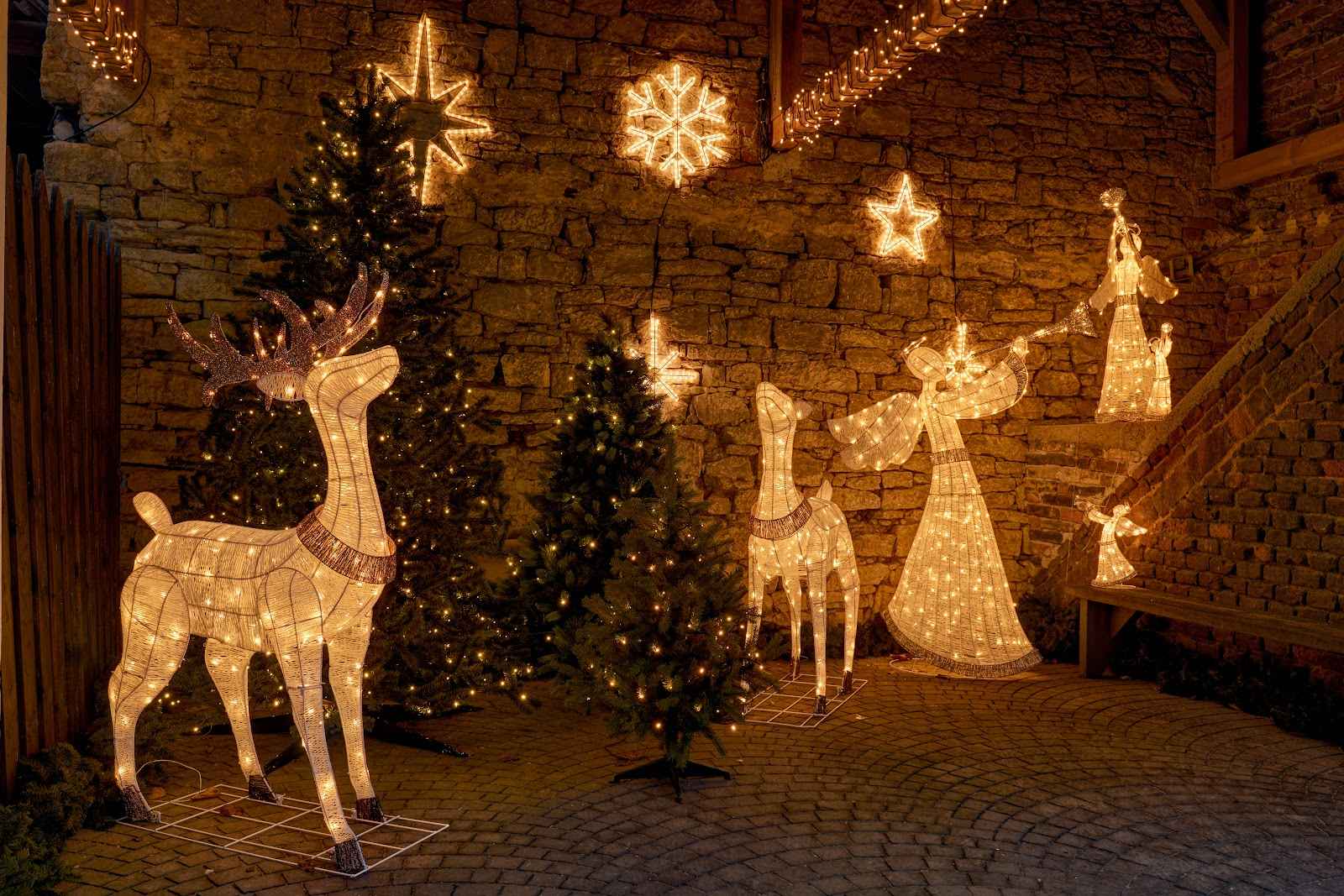 Figuras de Natal iluminadas, incluindo renas e anjos, dispostas num pátio exterior com paredes de pedra.